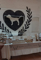 Indoor artisan market jewelry booth with organized ring and earring displays on a white table, butterfly-decorated banner, and a large black brick-wall mural of a dog silhouette inside a heart and laurel.
