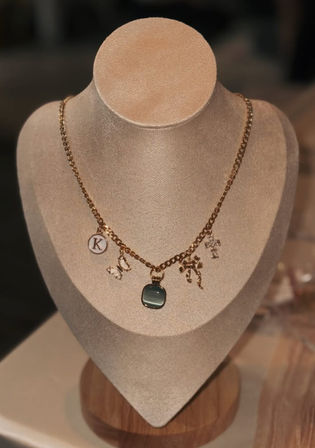 Gold charm necklace with a green square pendant and tiny bow, cross, butterfly and round initial disc charms displayed on a beige velvet bust in a boutique-style jewelry display.