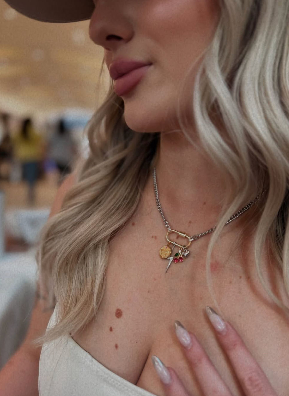 Close-up of a woman’s lower face and décolletage with wavy blonde hair, wearing a chunky silver chain necklace with mixed gold charms, metallic stiletto nails and a beige top in a blurred indoor setting.
