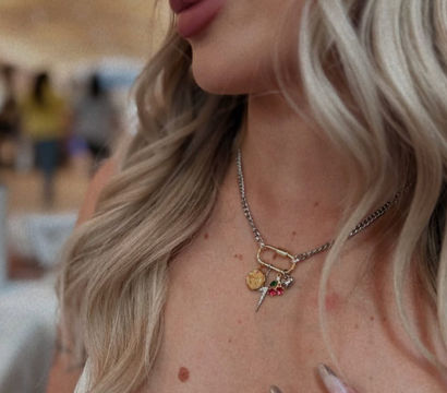 Close-up of a woman’s lower face and décolletage with wavy blonde hair, wearing a chunky silver chain necklace with mixed gold charms, metallic stiletto nails and a beige top in a blurred indoor setting.