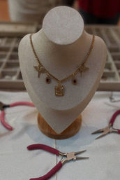 Gold charm necklace with dragonfly accents and red gemstone drops displayed on a beige jewelry bust, surrounded by pink-handled pliers on a craft table