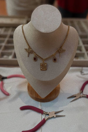 Gold charm necklace with dragonfly accents and red gemstone drops displayed on a beige jewelry bust, surrounded by pink-handled pliers on a craft table