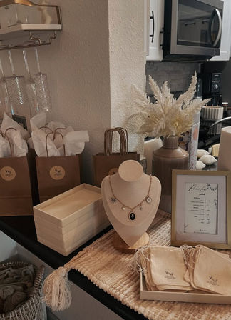 Neutral-toned jewelry display on a kitchen countertop — necklace on a fabric bust, stacked boxes, cloth pouches, brown gift bags, framed price list and a vase of pampas grass in a cozy boho setup.