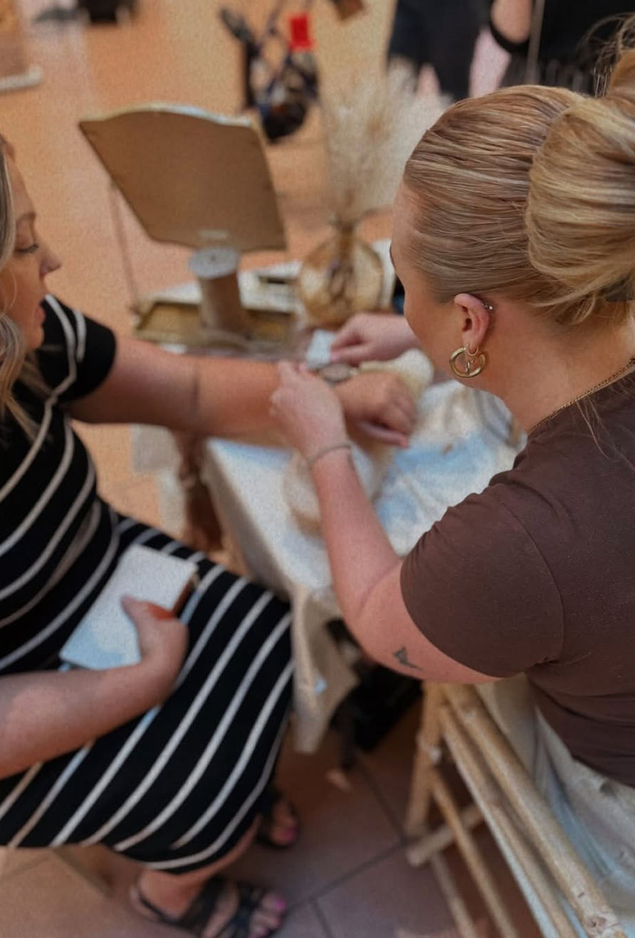 Two women at a boutique-style table—one with a neat bun fastening a bracelet on the other’s wrist, seated customer in a black-and-white striped dress with jewelry display and mirror in the background.