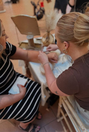 Two women at a boutique-style table—one with a neat bun fastening a bracelet on the other’s wrist, seated customer in a black-and-white striped dress with jewelry display and mirror in the background.