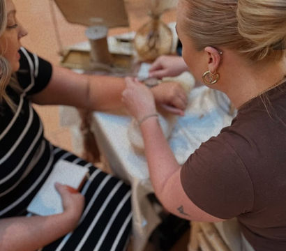 Two women at a boutique-style table—one with a neat bun fastening a bracelet on the other’s wrist, seated customer in a black-and-white striped dress with jewelry display and mirror in the background.