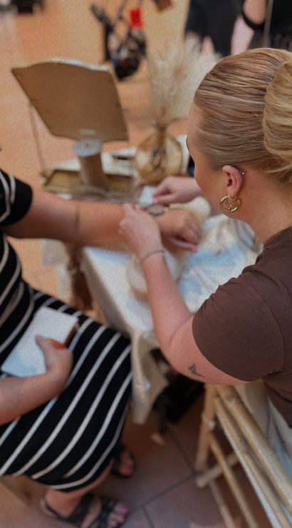 Two women at a boutique-style table—one with a neat bun fastening a bracelet on the other’s wrist, seated customer in a black-and-white striped dress with jewelry display and mirror in the background.