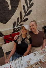 Two smiling women seated at an artisan jewelry stall with necklaces, rings and earrings displayed on a table in front of a painted brick mural.