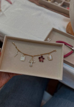 Hand holding a beige jewelry box with a gold charm bracelet on a craft table — five charms: lightning bolt, engraved rectangle tag, jeweled cross, red flower, and oval mother-of-pearl pendant; pliers visible in the background.