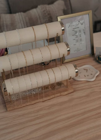 Three-tier beige and gold jewelry stand displaying delicate gold and silver chain necklaces on a light wood table, with a framed print and small dish of earrings in the background.