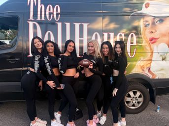 Six promo models in matching black athletic outfits smiling and posing with a football in front of a colorful promotional van featuring a large portrait, parked outdoors