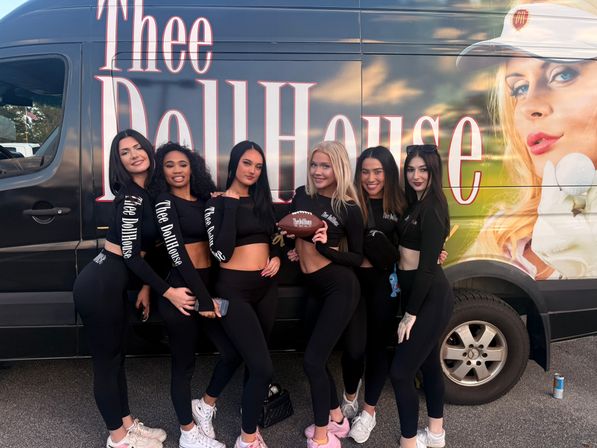 Six promo models in matching black athletic outfits smiling and posing with a football in front of a colorful promotional van featuring a large portrait, parked outdoors