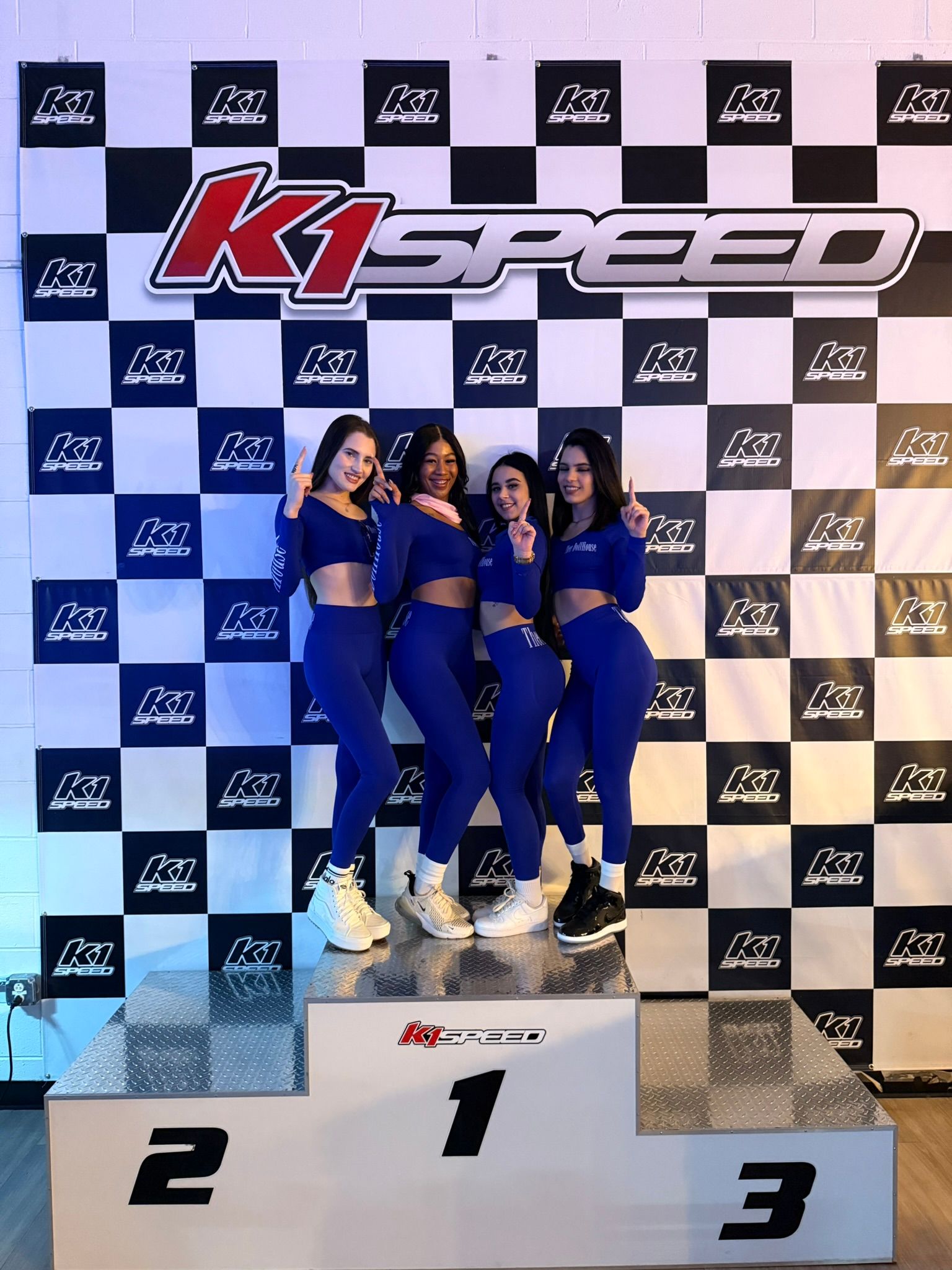 Four women in matching blue athletic outfits strike number-one poses on a winners podium at an indoor go-kart track, in front of a black-and-white checkered racing backdrop with a large logo