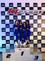 Four women in matching blue athletic outfits strike number-one poses on a winners podium at an indoor go-kart track, in front of a black-and-white checkered racing backdrop with a large logo