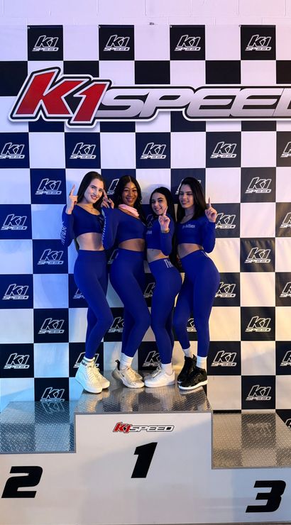 Four women in matching blue athletic outfits strike number-one poses on a winners podium at an indoor go-kart track, in front of a black-and-white checkered racing backdrop with a large logo