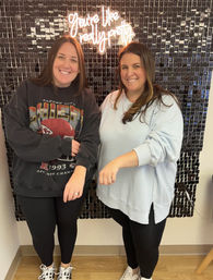 Two smiling friends standing in front of a black sequin photo wall with a neon sign reading "You're like really pretty," showing rings on their left hands; casual sweatshirts and leggings, indoor portrait.
