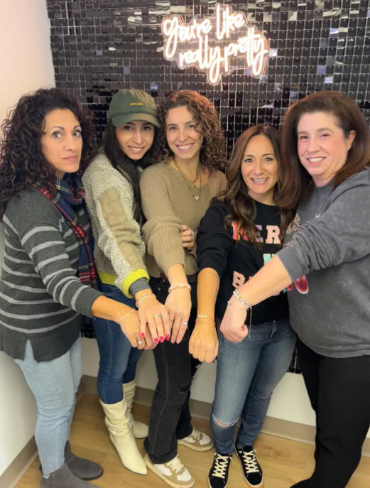 Five smiling women indoors show bracelets with joined hands, posing in front of a black tiled wall and neon sign reading 'you're like really pretty'.