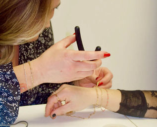 Close-up of hands at a jewelry counter measuring a wrist with a black sizing tool for a delicate gold bracelet; layered bracelets, rings, and red nail polish visible.