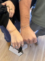 Two people showing wrists with delicate chain bracelets — gold on one wrist and silver on the other — hands with rings holding a small shopping bag on a light wood floor, jewelry shopping