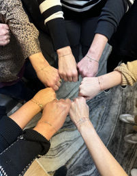 Top-down close-up of eight wrists forming a circle, fists touching to showcase delicate gold and beaded friendship bracelets on mixed skin tones and casual sleeves over a patterned rug.