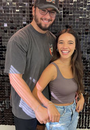 Smiling young couple holding hands in front of a shiny black sequin backdrop — man in baseball cap and tee, woman in cropped tank and ripped jeans in a playful candid pose.