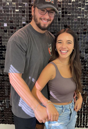 Smiling young couple holding hands in front of a shiny black sequin backdrop — man in baseball cap and tee, woman in cropped tank and ripped jeans in a playful candid pose.