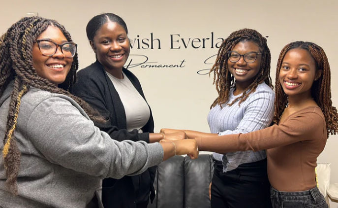 Four smiling women with braided hairstyles linking hands in a friendly circle inside a hair salon or beauty studio with neutral wall lettering and a couch.