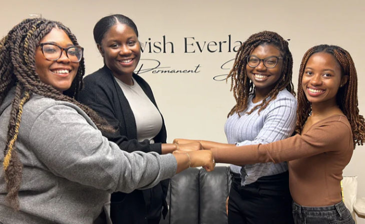 Four smiling women with braided hairstyles linking hands in a friendly circle inside a hair salon or beauty studio with neutral wall lettering and a couch.