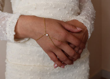 Close-up of folded hands over an ivory textured dress wearing a delicate gold hand-chain bracelet with a small square charm and pale pink manicure.