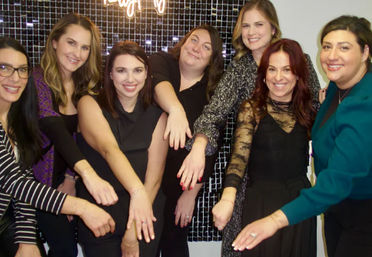 Seven smiling women posing indoors, playfully extending their hands to show bracelets and rings in a group photo against a black tiled backdrop