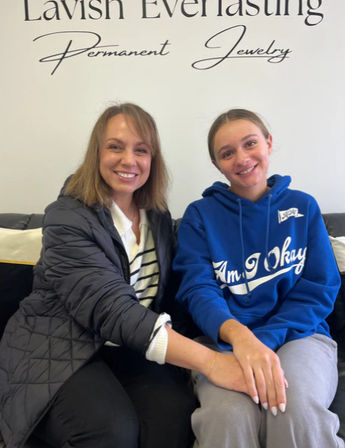 Two smiling customers seated on a black couch holding hands inside a permanent jewelry studio, one wearing a blue hoodie and gray sweatpants, the other in a black quilted jacket and striped top with a white wall sign behind them.