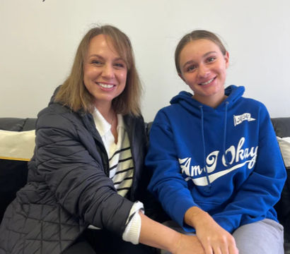 Two smiling women sitting on a couch holding hands inside a bright permanent-jewelry studio with stylish wall lettering