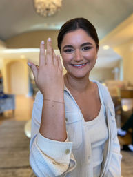 Smiling woman in a bright home interior holding up her left hand to display a ring, wearing a cream textured jacket and tank top with a chandelier-lit background.