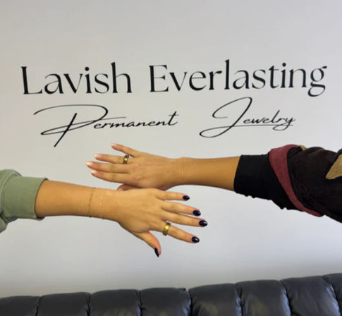 Two people extending hands to display matching delicate gold permanent bracelets and rings with black nail polish, posed in front of a script wall sign inside a jewelry studio above a black leather couch.
