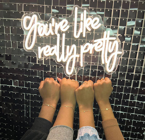Four raised wrists with varying skin tones wearing delicate bracelets beneath a white neon script sign reading 'You're like really pretty' on a black sequin backdrop.