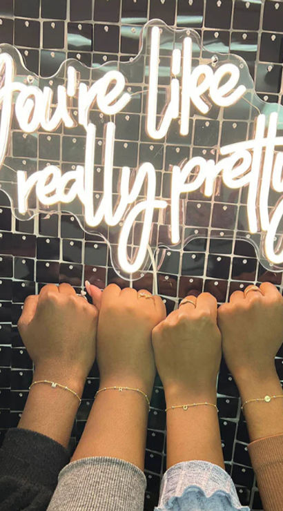Four raised wrists with varying skin tones wearing delicate bracelets beneath a white neon script sign reading 'You're like really pretty' on a black sequin backdrop.
