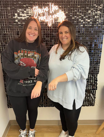 Two smiling friends pose in front of a black sequin photo wall with a neon sign reading 'You're like really pretty,' both showing rings on their left hands while wearing casual sweatshirts, leggings, and sneakers.