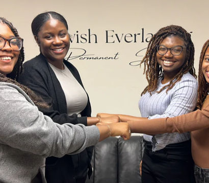 Four smiling women link hands in a friendly team pose inside an office studio with decorative wall lettering, wearing casual outfits and braided hairstyles.