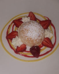 Powdered-sugar–topped round pastry on a white plate, surrounded by sliced strawberries, whipped cream rosettes and concentric red berry and yellow citrus sauce rings — plated strawberry dessert.