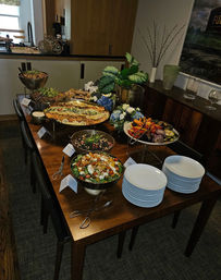 Indoor dining-room buffet on a wooden table with stacked white plates, mixed salads, roasted vegetables, bread, serving tongs and a leafy centerpiece.