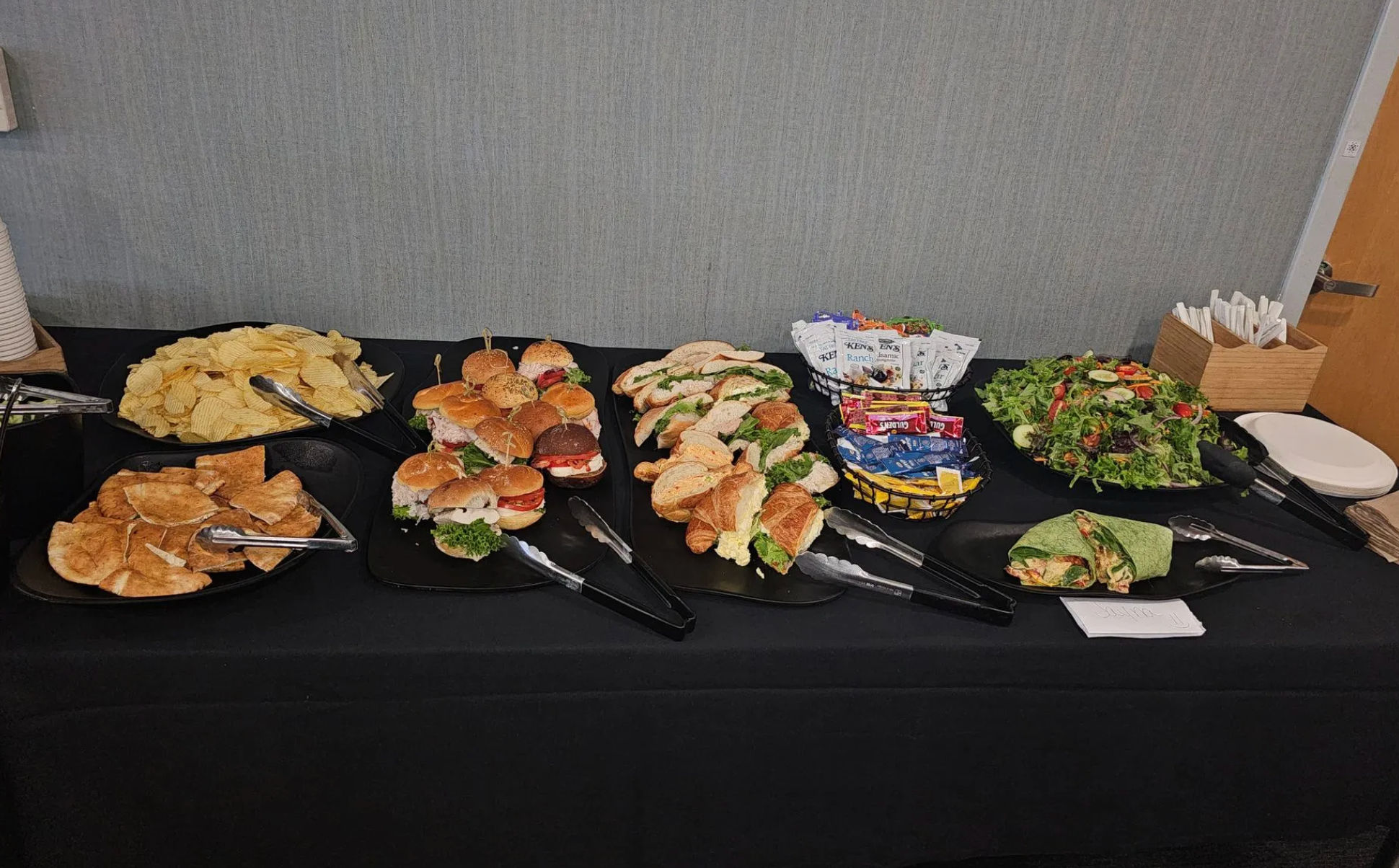 Catered office lunch buffet on a black tablecloth with assorted mini sandwiches and croissant rolls, wraps, potato and pita chips, mixed green salad, snack packets and disposable plates and utensils.