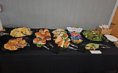 Catered office lunch buffet on a black tablecloth with assorted mini sandwiches and croissant rolls, wraps, potato and pita chips, mixed green salad, snack packets and disposable plates and utensils.