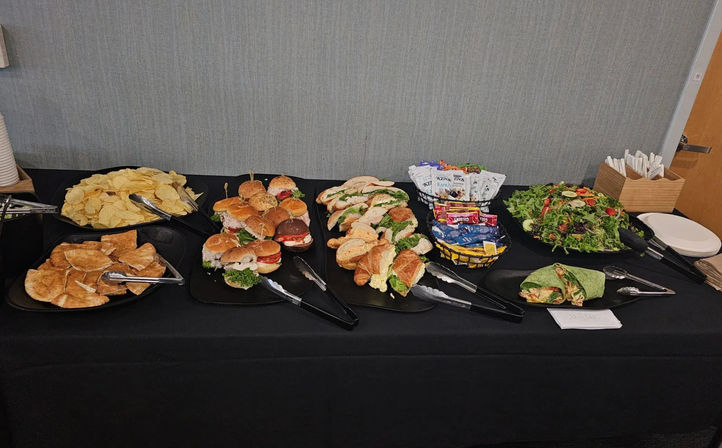 Catered office lunch buffet on a black tablecloth with assorted mini sandwiches and croissant rolls, wraps, potato and pita chips, mixed green salad, snack packets and disposable plates and utensils.