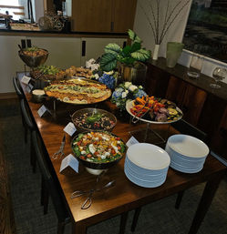 Colorful indoor catered buffet on a wooden dining table with stacked white plates, mixed greens and tomato salad, grain and bean bowls, lemon-garnished paella, roasted vegetables, serving tongs and a floral centerpiece in a modern dining room.