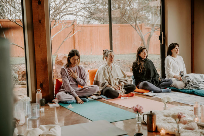 Four participants in a cozy sunlit meditation class seated on colorful yoga mats and cushions near a large window overlooking a fenced backyard