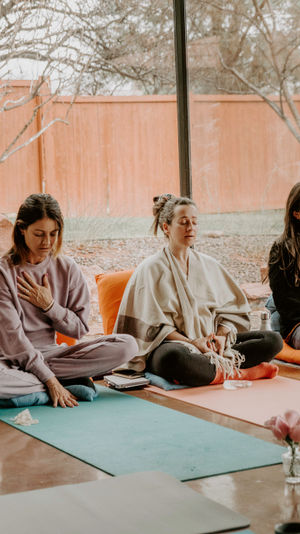 Four participants in a cozy sunlit meditation class seated on colorful yoga mats and cushions near a large window overlooking a fenced backyard