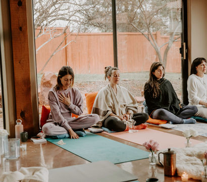 Four participants in a cozy sunlit meditation class seated on colorful yoga mats and cushions near a large window overlooking a fenced backyard