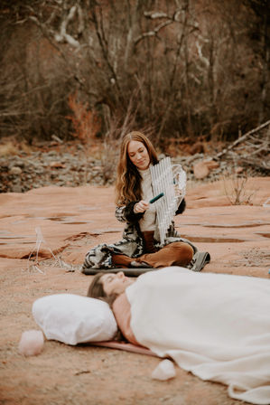 Sound healing on red sandstone: woman in a red-rock desert canyon plays metal chimes with a mallet beside a resting person.