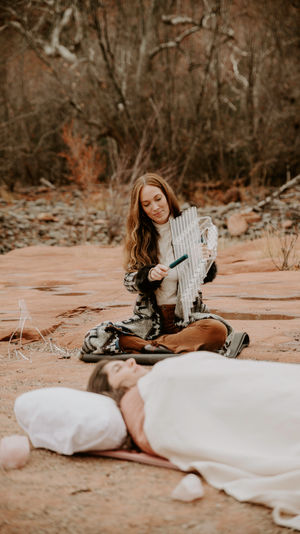 Sound healing on red sandstone: woman in a red-rock desert canyon plays metal chimes with a mallet beside a resting person.