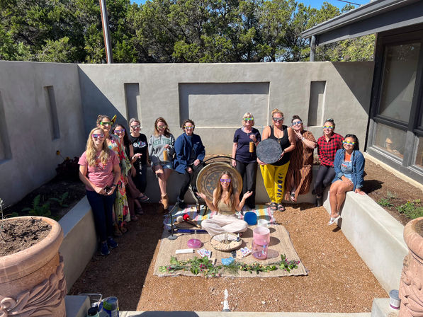 Sunlit outdoor stucco courtyard with about a dozen people wearing colorful eclipse glasses, gathered around a seated facilitator and a sound-healing setup with a gong, drum and crystal singing bowls on a woven mat, trees visible above the wall.